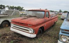 an old red truck parked in a field next to other cars and trucks with rust on the hoods