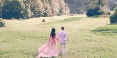 a man and woman are walking through the grass in front of some trees on a hill