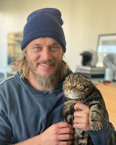 a man holding a cat in his arms while wearing a blue beanie and smiling at the camera