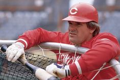 a baseball player leaning on the net with his bat in hand and wearing a red uniform