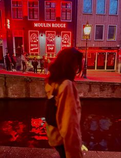 a woman walking down a street next to a body of water in front of a red lit building