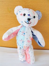 a white teddy bear with pink and blue flowers on it's chest, sitting against a wooden wall