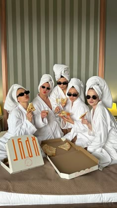 four women in white robes eating pizza and drinking wine on a bed with striped wallpaper