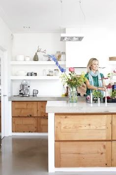 a woman standing in a kitchen next to a counter with flowers on top of it