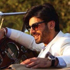 a man wearing sunglasses and a white suit sitting in a car with his hand on the steering wheel