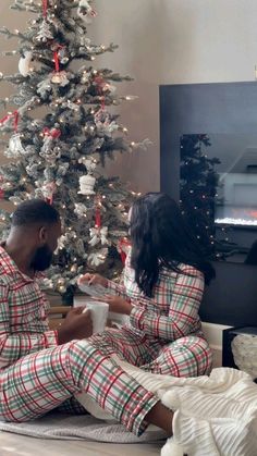 a man and woman sitting on the floor in matching pajamas next to a christmas tree