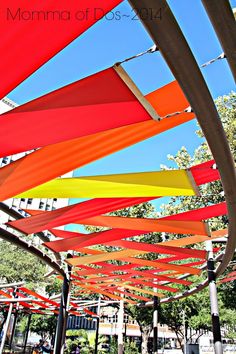 an array of colorful umbrellas hanging from the ceiling in front of trees and buildings