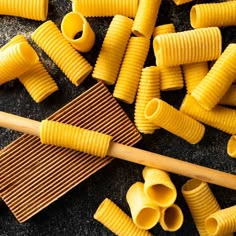 some yellow pasta is next to a wooden spatula on a black surface with other food items