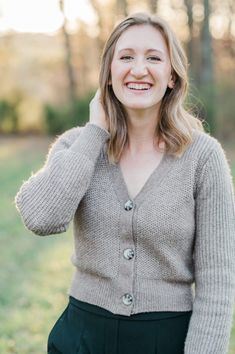 a woman in a cardigan smiles as she poses for the camera with her hand on her ear