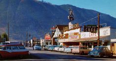 an old photo of cars parked on the street in front of shops and businesses with mountains in the background