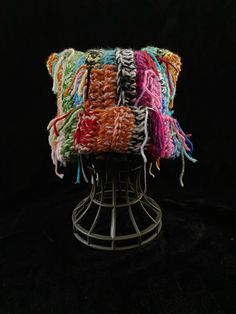 a multicolored knitted pillow sitting on top of a metal stand against a black background