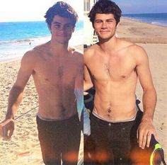 two men standing next to each other at the beach and one holding a surfboard