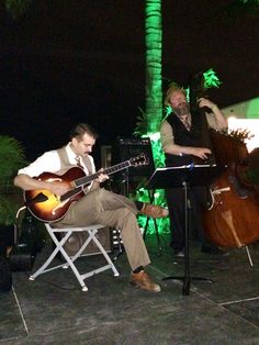 two men are playing music on the patio at night with palm trees in the background