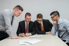 three men in business attire look at papers on a table while another man looks down