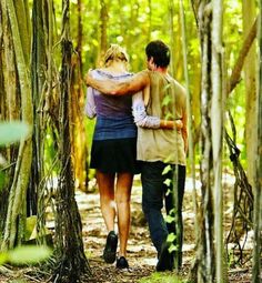 two people walking through the woods with their arms around each other, hugging and embracing