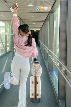 a woman in white pants and pink sweater pulling luggage down a hallway with her hand up