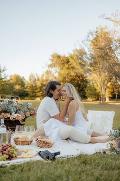 a man and woman sitting on a blanket in the grass with food, drinks and wine