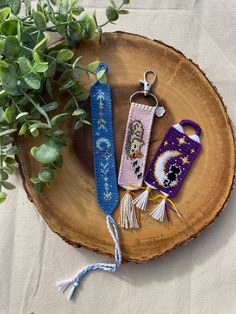 two cross stitch keychains sitting on top of a piece of wood next to a plant