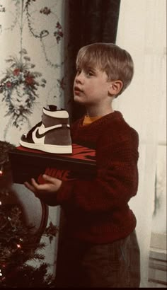 a young boy holding a shoe box in front of a christmas tree