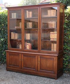 an old wooden bookcase with many books on the top and bottom shelves, in front of a hedge