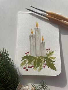 two white candles with holly and berries on them are sitting next to some paintbrushes