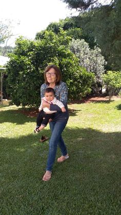 a woman holding a small child in her arms on the grass with trees and bushes behind her