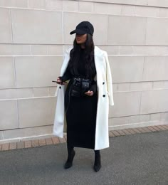 a woman standing in front of a brick wall wearing a white coat and black hat