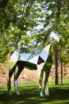 a horse made out of mirrored glass in the grass with trees and bushes behind it