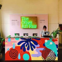 a woman sitting at the front desk of an art fair with potted plants behind her