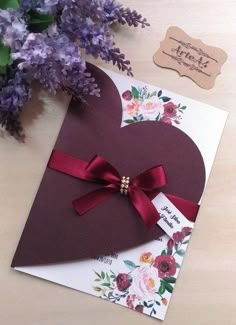 a heart shaped card with a red ribbon on it next to purple flowers and a tag