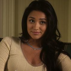 a woman with long dark hair wearing a tan shirt and blue beaded necklace sitting in front of a window