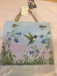 a painting of a hummingbird flying over blue flowers on a white sheeted surface