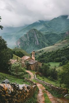 an old church sits in the middle of a valley with green mountains behind it and a dirt path leading up to it