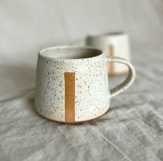 two coffee mugs sitting on top of a bed next to each other, one with an orange stripe