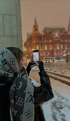 a woman standing in front of a building taking a photo with her cell phone while it is snowing