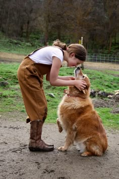 a woman petting a large brown dog on top of a dirt field with trees in the background