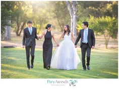 three people in formal wear holding hands and walking through the grass with trees behind them