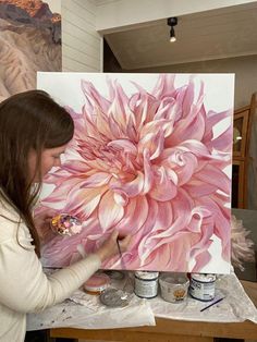 a woman painting a large pink flower on a canvas with watercolors in front of her