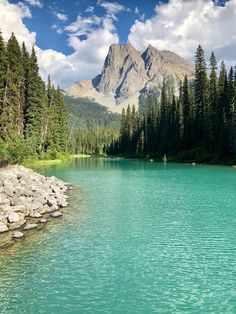 a lake surrounded by trees and mountains under a blue sky with puffy white clouds