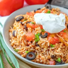 a white bowl filled with mexican rice and black olives next to tomatoes, sour cream on top