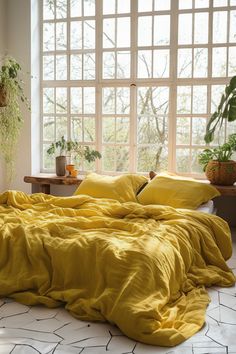 a bed with yellow sheets and pillows in front of a window filled with potted plants