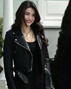 a woman wearing a black leather jacket is smiling at the camera while standing in front of a white building