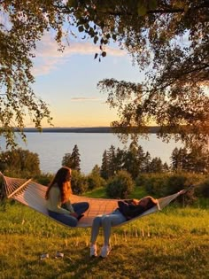 two people are sitting in a hammock on the grass by the water at sunset