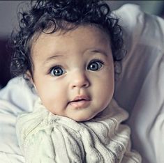 a baby with curly hair wearing a white sweater and looking at the camera while laying on a bed