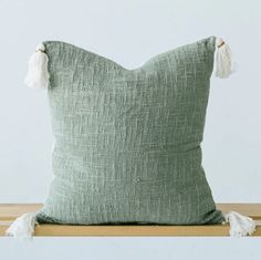 a green pillow with white tassels on top of a wooden table next to a wall