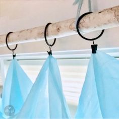 three blue bags hanging from a wooden rod in front of a window with white curtains