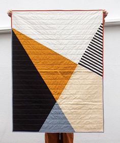 a person holding up a large quilt in front of a white wall with an orange, black and yellow design on it