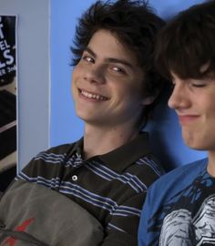 two young men sitting next to each other in front of a wall with posters on it