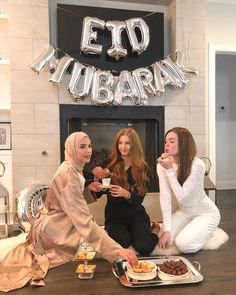 three women are sitting on the floor eating food and drinking tea in front of a sign that says eid mubaran