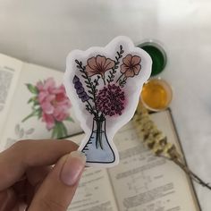 a hand holding a sticker with flowers in a vase on top of an open book